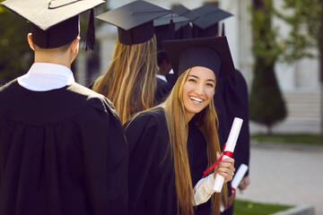 Wall Mural - Happy cute young graduate girl looks back with smile during graduation event for college students and holds certificate of graduation stands in line among peers on campus of university