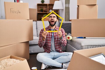 Wall Mural - Middle east man with beard sitting on the floor at new home holding ruler angry and mad screaming frustrated and furious, shouting with anger looking up.