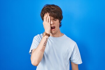 Sticker - Hispanic young man standing over blue background yawning tired covering half face, eye and mouth with hand. face hurts in pain.