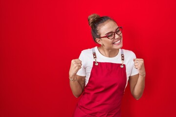 Wall Mural - Young hispanic woman wearing waitress apron over red background very happy and excited doing winner gesture with arms raised, smiling and screaming for success. celebration concept.