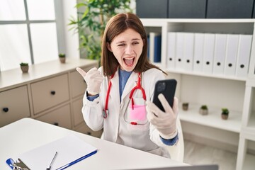 Poster - Young doctor woman doing video call with smartphone pointing thumb up to the side smiling happy with open mouth