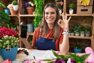 Sticker - Hispanic young woman working at florist shop showing smartphone screen doing ok sign with fingers, smiling friendly gesturing excellent symbol