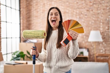 Poster - Young brunette woman holding roller painter and paint samples celebrating crazy and amazed for success with open eyes screaming excited.