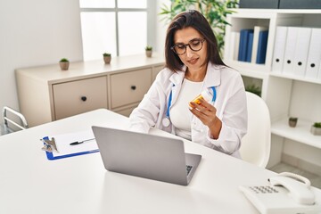 Poster - Young hispanic woman wearing doctor uniform holding pills working at clinic