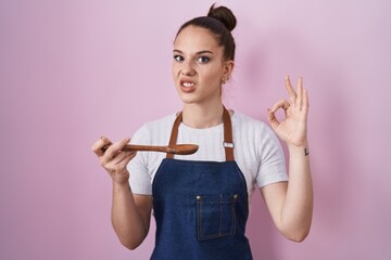 Poster - Young hispanic girl wearing professional cook apron holding wood spoon clueless and confused expression. doubt concept.