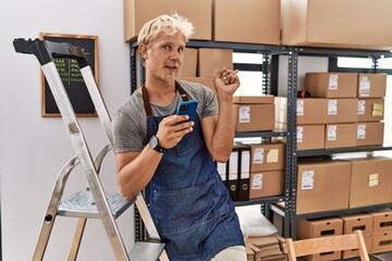 Canvas Print - Young blond man using smartphone working at storehouse with a big smile on face, pointing with hand finger to the side looking at the camera.