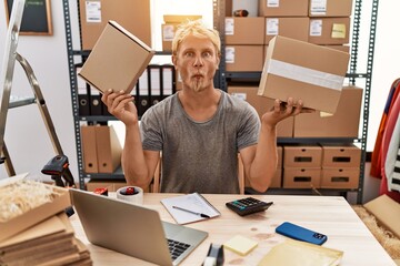 Sticker - Young blond man holding packages working at online shop making fish face with mouth and squinting eyes, crazy and comical.