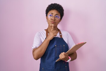 Canvas Print - Young african american woman wearing professional waitress apron holding clipboard thinking concentrated about doubt with finger on chin and looking up wondering