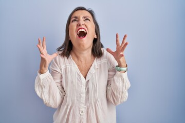 Sticker - Middle age hispanic woman standing over blue background crazy and mad shouting and yelling with aggressive expression and arms raised. frustration concept.