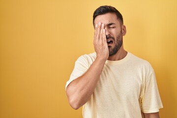 Poster - Handsome hispanic man standing over yellow background yawning tired covering half face, eye and mouth with hand. face hurts in pain.