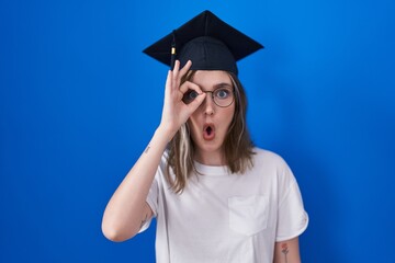 Wall Mural - Blonde caucasian woman wearing graduation cap doing ok gesture shocked with surprised face, eye looking through fingers. unbelieving expression.