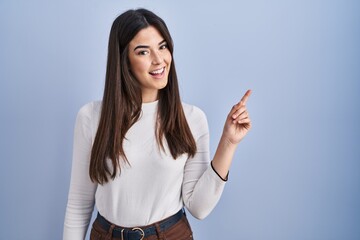 Poster - Young brunette woman standing over blue background with a big smile on face, pointing with hand finger to the side looking at the camera.