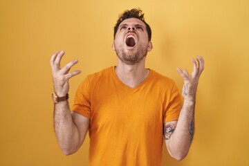 Poster - Young hispanic man standing over yellow background crazy and mad shouting and yelling with aggressive expression and arms raised. frustration concept.