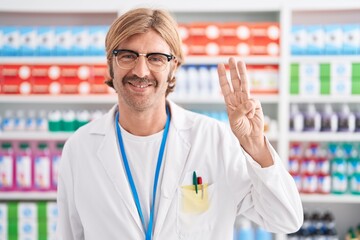 Canvas Print - Caucasian man with mustache working at pharmacy drugstore showing and pointing up with fingers number three while smiling confident and happy.