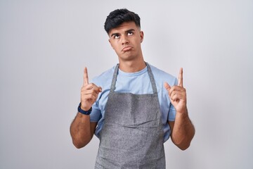 Sticker - Hispanic young man wearing apron over white background pointing up looking sad and upset, indicating direction with fingers, unhappy and depressed.