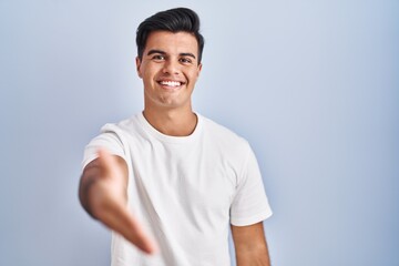 Poster - Hispanic man standing over blue background smiling friendly offering handshake as greeting and welcoming. successful business.