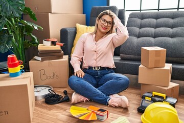 Canvas Print - Young hispanic woman moving to a new home sitting on the floor smiling confident touching hair with hand up gesture, posing attractive and fashionable