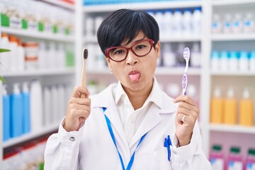 Sticker - Young asian woman with short hair doing toothbrush comparative at pharmacy sticking tongue out happy with funny expression.