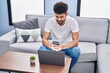 © Krakenimages.com - Young arab man using smartphone and laptop sitting on sofa at home
