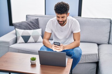 Canvas Print - Young arab man using smartphone and laptop sitting on sofa at home