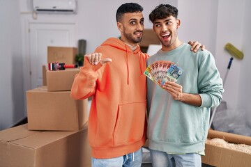 Poster - Young hispanic gay couple moving to a new home holding banknotes pointing thumb up to the side smiling happy with open mouth