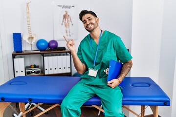 Poster - Young physiotherapist man working at pain recovery clinic with a big smile on face, pointing with hand finger to the side looking at the camera.