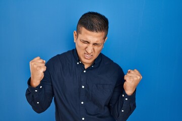 Sticker - Hispanic young man standing over blue background very happy and excited doing winner gesture with arms raised, smiling and screaming for success. celebration concept.