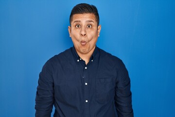 Canvas Print - Hispanic young man standing over blue background making fish face with lips, crazy and comical gesture. funny expression.