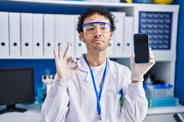 Canvas Print - Hispanic man working at scientist laboratory showing smartphone screen doing ok sign with fingers, smiling friendly gesturing excellent symbol