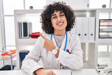 Poster - Young brunette woman with curly hair wearing doctor uniform and stethoscope smiling cheerful pointing with hand and finger up to the side