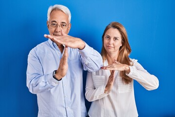 Wall Mural - Middle age hispanic couple standing over blue background doing time out gesture with hands, frustrated and serious face