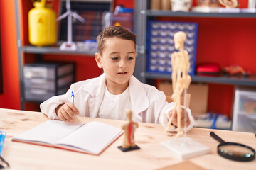 Poster - Adorable hispanic toddler student writing on notebook looking skeleton at classroom