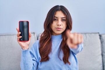 Sticker - Hispanic young woman holding smartphone showing blank screen pointing with finger to the camera and to you, confident gesture looking serious