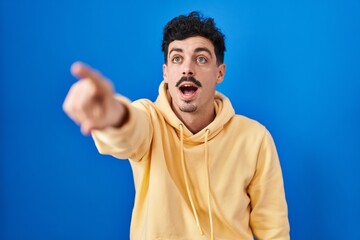 Wall Mural - Hispanic man standing over blue background pointing with finger surprised ahead, open mouth amazed expression, something on the front