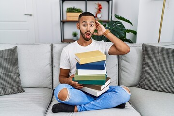 Poster - African american young man holding a pile of books sitting on the sofa crazy and scared with hands on head, afraid and surprised of shock with open mouth