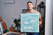 © Krakenimages.com - Young hispanic painter man holding canvas smiling with a happy and cool smile on face. showing teeth.