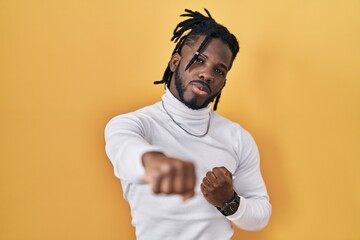 Poster - African man with dreadlocks wearing turtleneck sweater over yellow background punching fist to fight, aggressive and angry attack, threat and violence