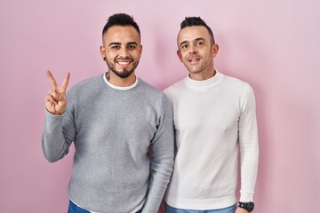 Sticker - Homosexual couple standing over pink background showing and pointing up with fingers number two while smiling confident and happy.