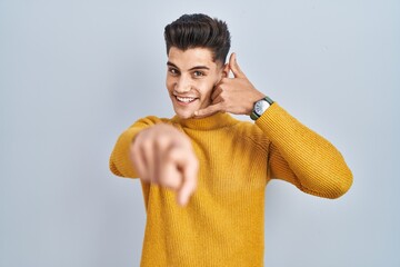 Wall Mural - Young hispanic man standing over blue background smiling doing talking on the telephone gesture and pointing to you. call me.