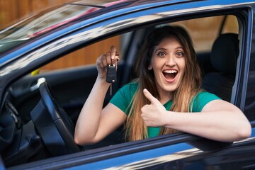 Sticker - Young brunette woman holding key of new car pointing thumb up to the side smiling happy with open mouth