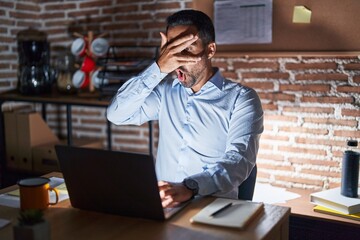 Canvas Print - Hispanic man with beard working at the office at night peeking in shock covering face and eyes with hand, looking through fingers with embarrassed expression.