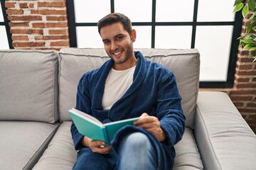 Wall Mural - Young hispanic man wearing bathrobe reading book at home