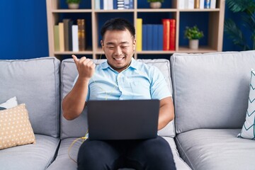 Sticker - Chinese young man using computer laptop sitting on the sofa pointing thumb up to the side smiling happy with open mouth