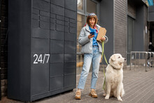 Young Woman Uses Phone While Receiving A Parcel From Automatic Post Office Machine During A Walk With Her Dog In City. Concept Of Modern Technologies In Delivery Services And Lifestyle