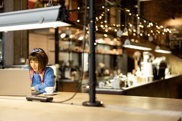 Wall Mural - Young stylish woman works on laptop while sitting with a coffee drink at modern cafe. Concept of remote work from public place, digital freelance and modern lifestyle