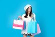 Beautiful smiling asian woman, white skin with long hair, casual wear, wearing a hat, she is holding several shopping bags of various colors, with a smartphone, on a blue background in a studio.