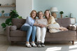 Cheerful loving old grandmother, mature mother, young adult daughter woman sitting together on comfortable couch in modern home living room interior, looking at camera, smiling. Full length shot