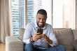 © fizkes - Handsome single African guy sit on sofa at modern apartment with cell phone, looks at smartphone screen read sms, message, chat with friend online, enjoy e-dating services spend leisure on internet