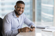 © fizkes - Portrait of happy successful 35s African businessman sit at workplace desk with smartphone smile looking at camera. Modern workspace, wireless technologies, new professional mobile application usage