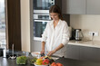 © fizkes - Beautiful slim woman cooking in cozy domestic kitchen, cuts fresh avocado for vegetarian salad smile enjoys cookery process. Homemade food, vegetables preparation, weight loss, healthy dietary recipes